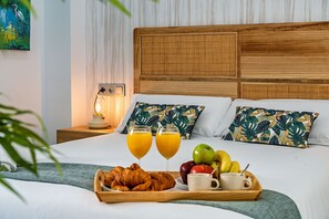 Ein ordentlich bezogenes Bett mit Holz-Kopfstück, zwei Kissen mit tropischem Blattmuster, ein Nachttisch mit Lampe und eine Ablage mit Croissants, Obst und Orangensaft.