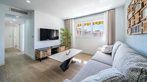 Ein modernes Wohnzimmer mit Flachbildfernseher, Couchtisch aus Holz, grauem Sofa und einem großen Fenster mit Vorhängen.