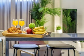 A breakfast tray with croissants, an apple, bananas, and orange juice on a wooden table in a modern kitchen with a TV and plants in the background.
