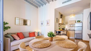 Una sala moderna con una mesa redonda de madera, sillas con cojines azules, un sofá beige con cojines de colores, un camino de mesa tejido y una planta en maceta.