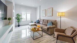 Ein modernes Wohnzimmer mit einer Couch, einem Couchtisch und einer Stehlampe. Zwei Fenster und ein an der Wand montierter Fernseher sind zu sehen.