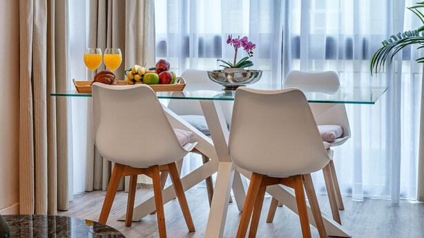 Un comedor moderno con una mesa de vidrio, sillas blancas y una bandeja de frutas con vasos de jugo.