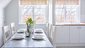 Un comedor con una mesa dispuesta para cuatro personas, sillas blancas, un jarrón con flores y una ventana con persianas.
