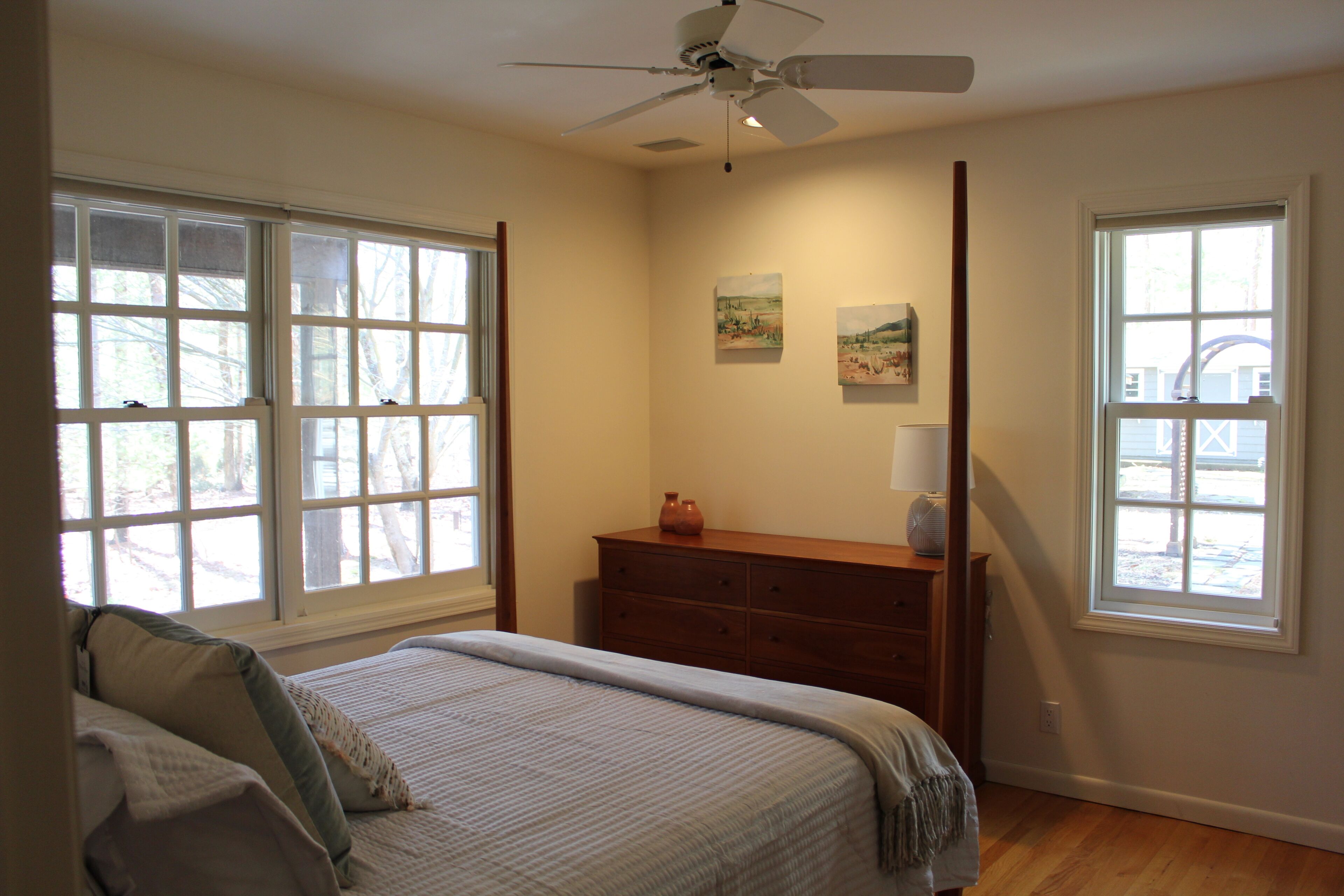 Ein Schlafzimmer mit Bett, Schminktisch, zwei Fenstern, Deckenventilator und Kunstwerken an der Wand.