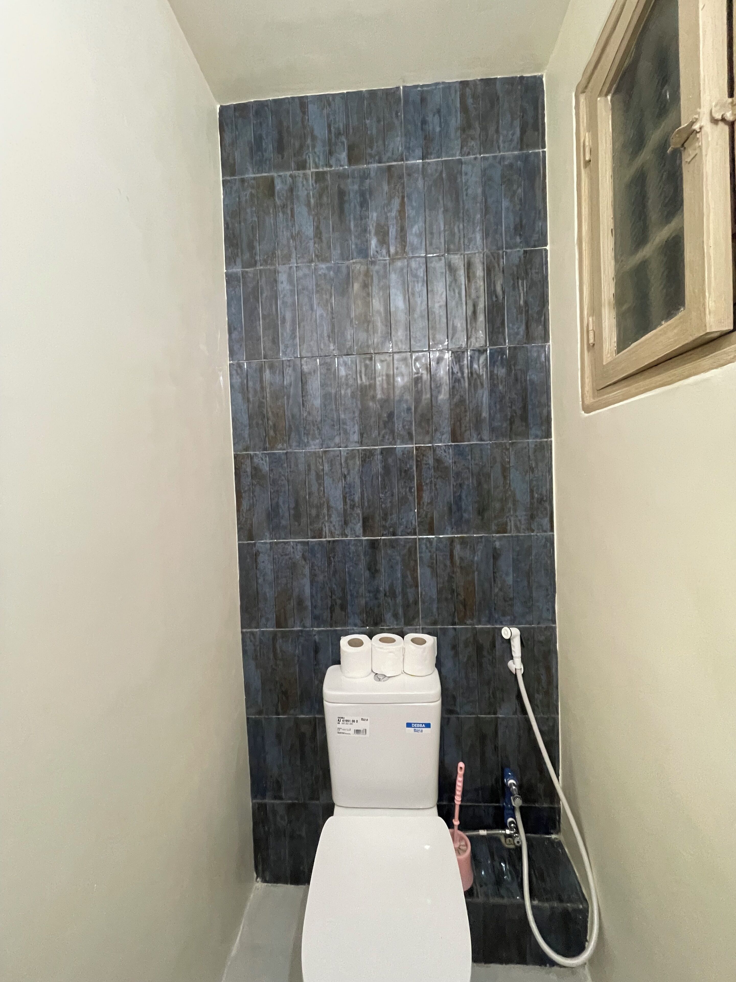 Un baño con inodoro, pared de azulejos oscuros y una ventana.