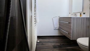 Moderni kylpyhuone, jossa on tumma puulattia, valkoinen wc-istuin, puinen allaskaappi peilillä ja pyyhe, joka on ripustettu seinälle.