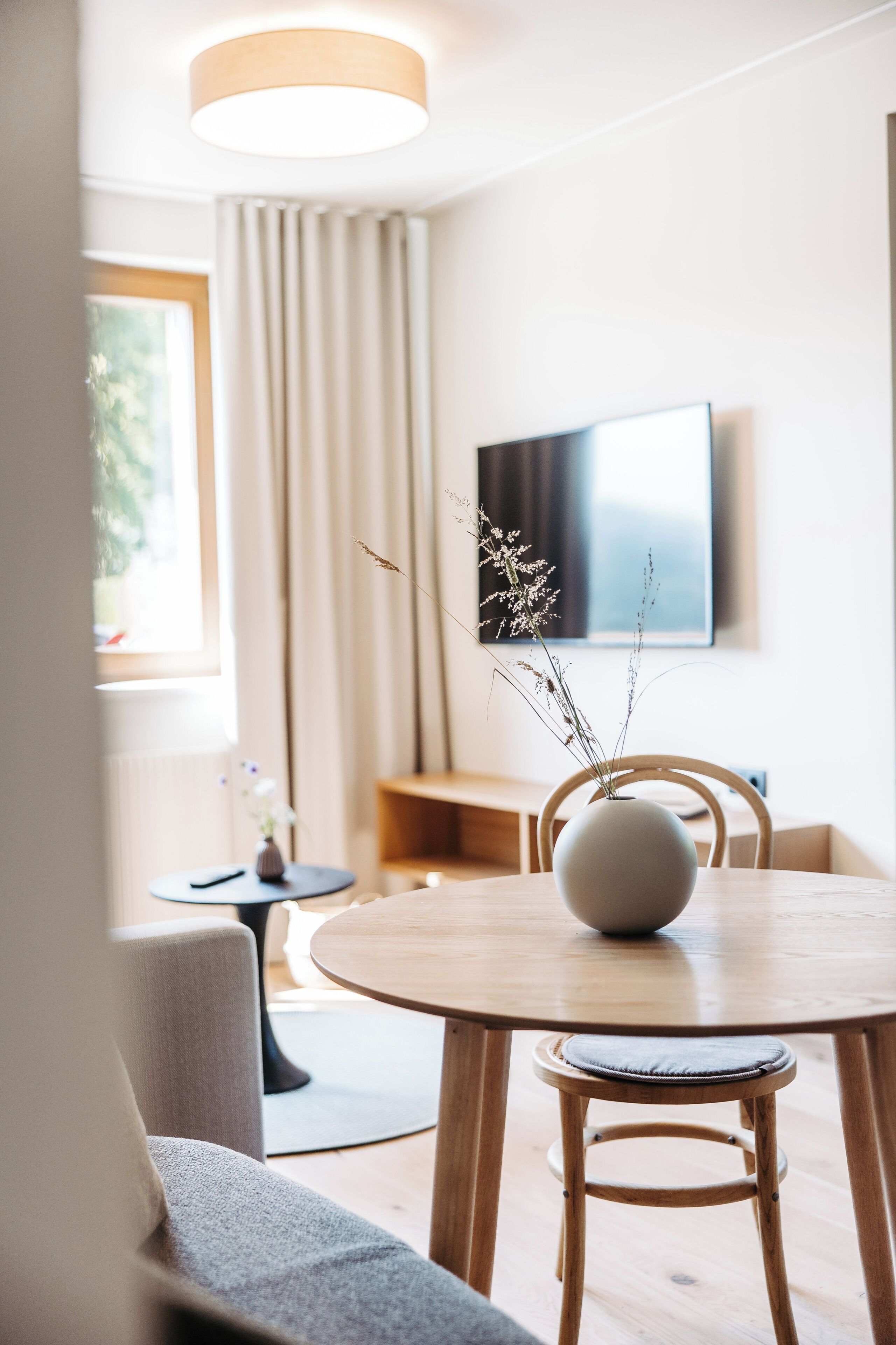 A modern dining area with a round wooden table, a vase with dried flowers, and a television mounted on the wall.