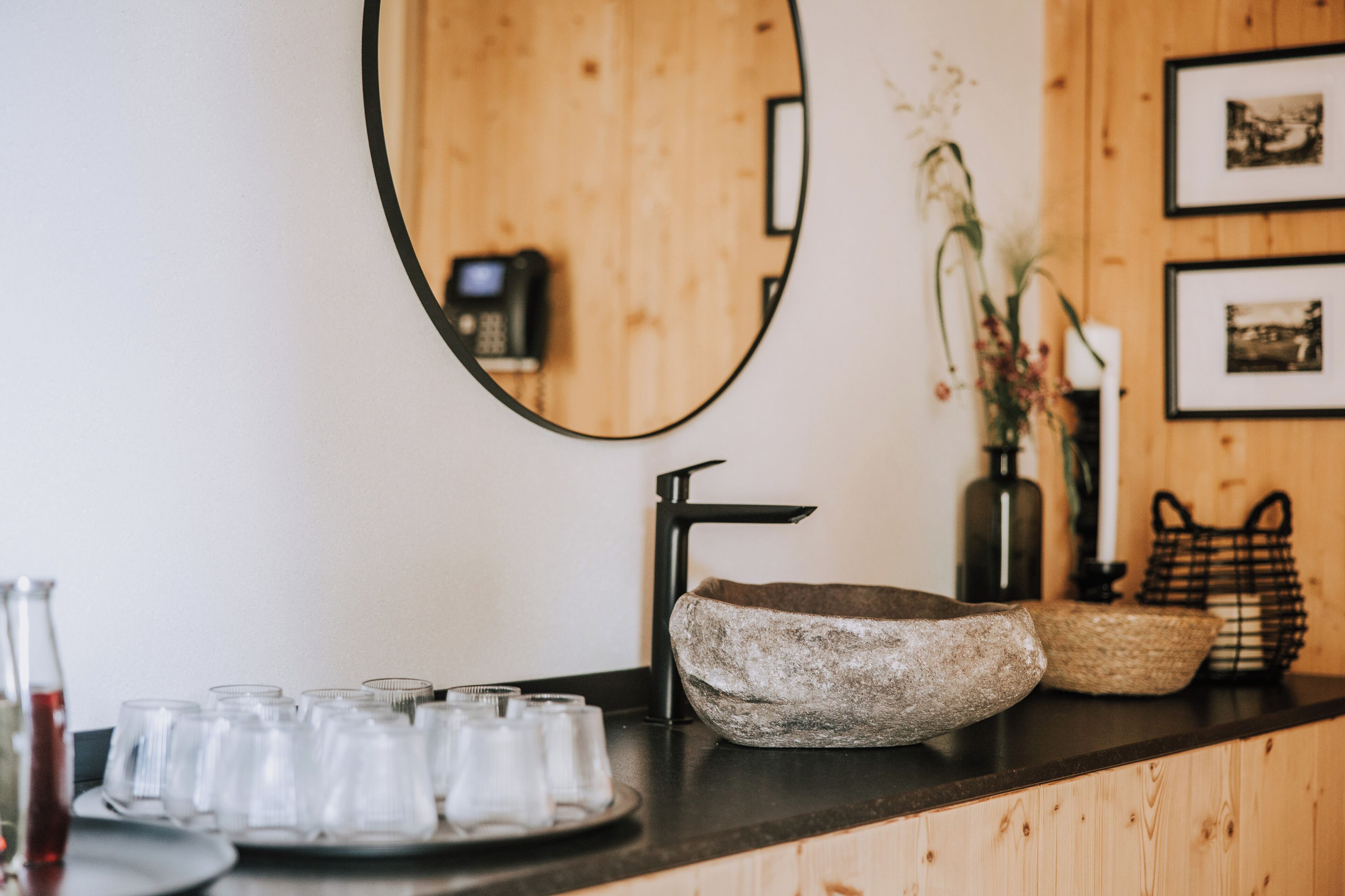 A bathroom with a wooden vanity, a round mirror, a black faucet, and a tray of glasses.