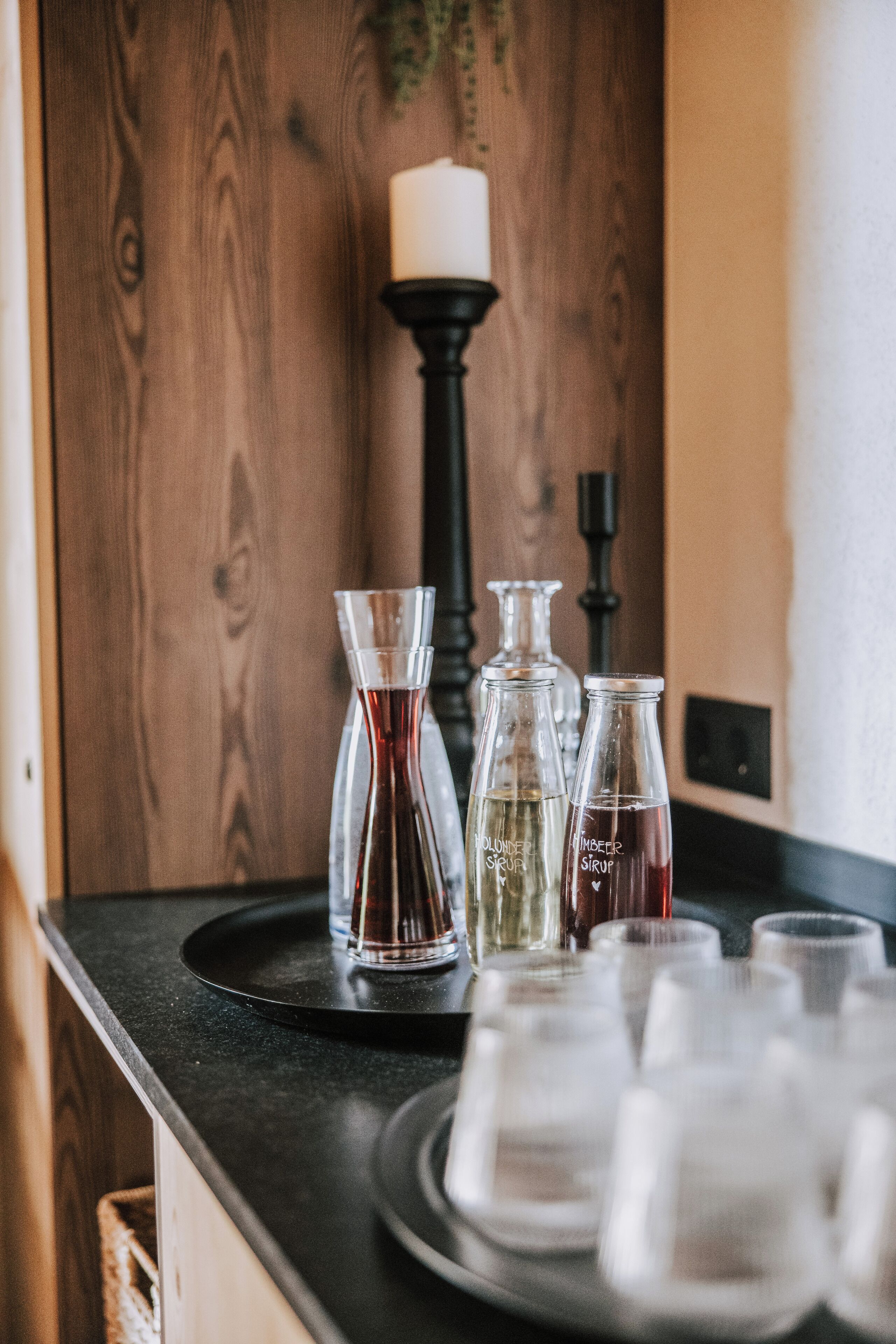 Una encimera de cocina con una bandeja negra que contiene una jarra de vidrio, dos botellas de vidrio etiquetadas como "VODKELDER SEIF" y "TIMBER SIRUP", y varios vasos vacíos.