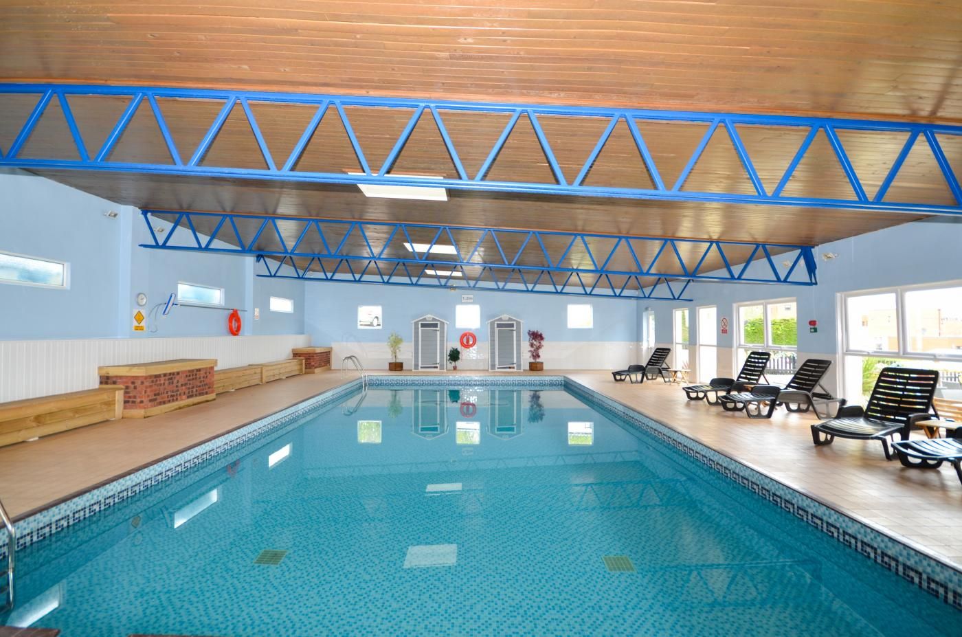 Une piscine intérieure avec un sol carrelé de bleu, un plafond en bois avec des poutres bleues et de grandes fenêtres baignées de lumière.