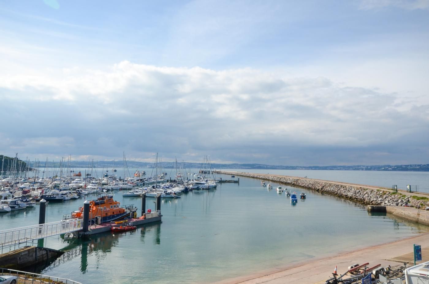 Un port de plaisance où sont amarrés de nombreux bateaux, une jetée et une mer calme sous un ciel nuageux.