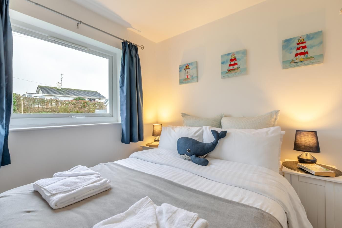 A hotel room with a bed, two bedside lamps, a window with curtains, and three paintings on the wall.