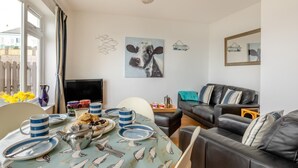 A living room with a dining table set for two, a black leather sofa, a TV, and a painting of a cow on the wall. - 2 Bed in Brixham (oc-f28884) (Brixham)