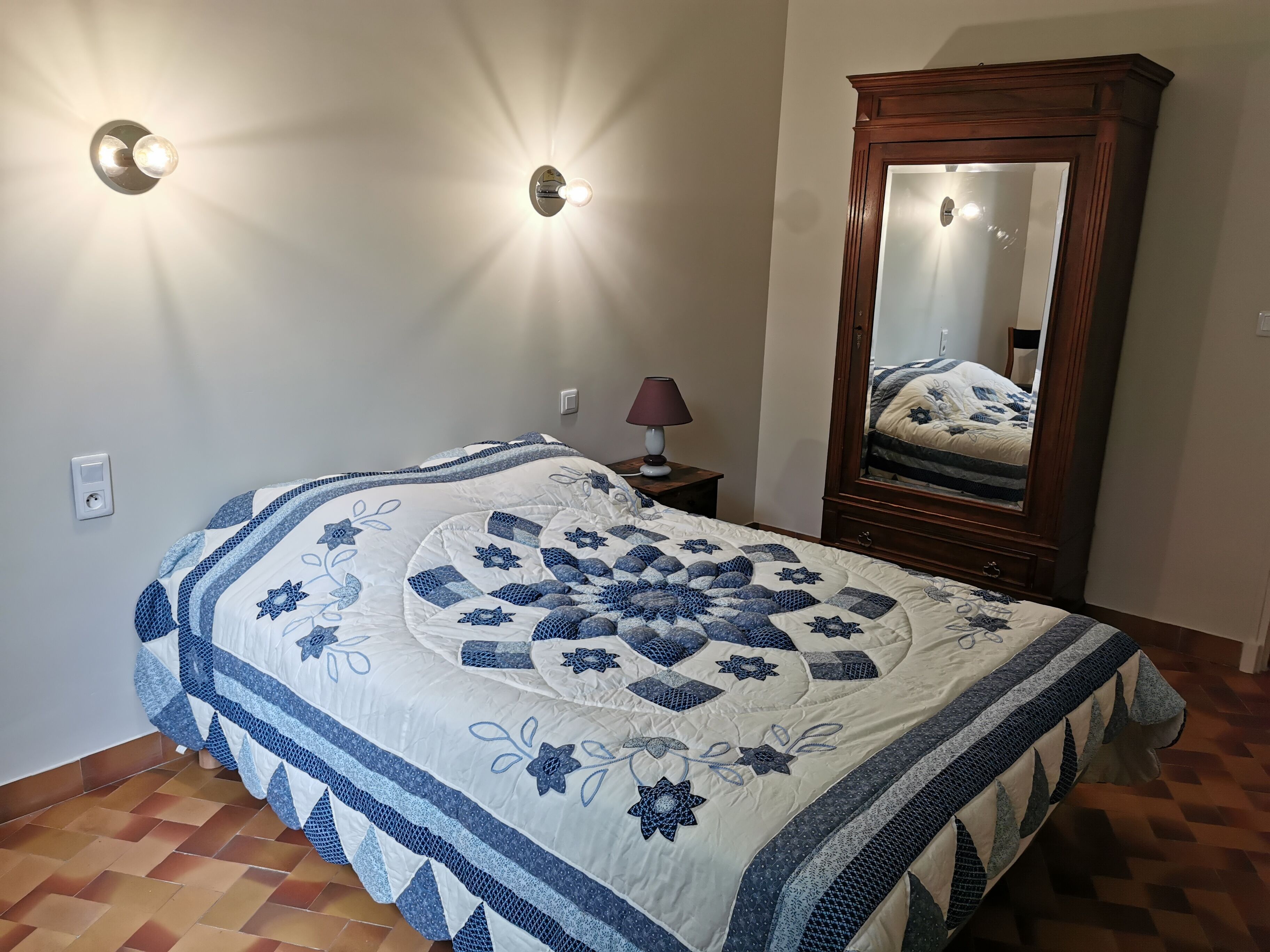 A bedroom with a bed, a nightstand, a lamp, a mirror, and a wall-mounted light fixture.