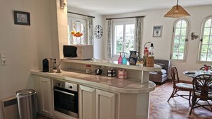 Een keuken met een centraal kookeiland, een magnetron, een koffiezetapparaat en een prullenbak. Er is een eethoek met een tafel en stoelen.