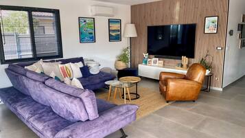 A modern living room with a purple sofa, a brown leather chair, a glass coffee table, and a flat-screen TV mounted on a wooden wall.