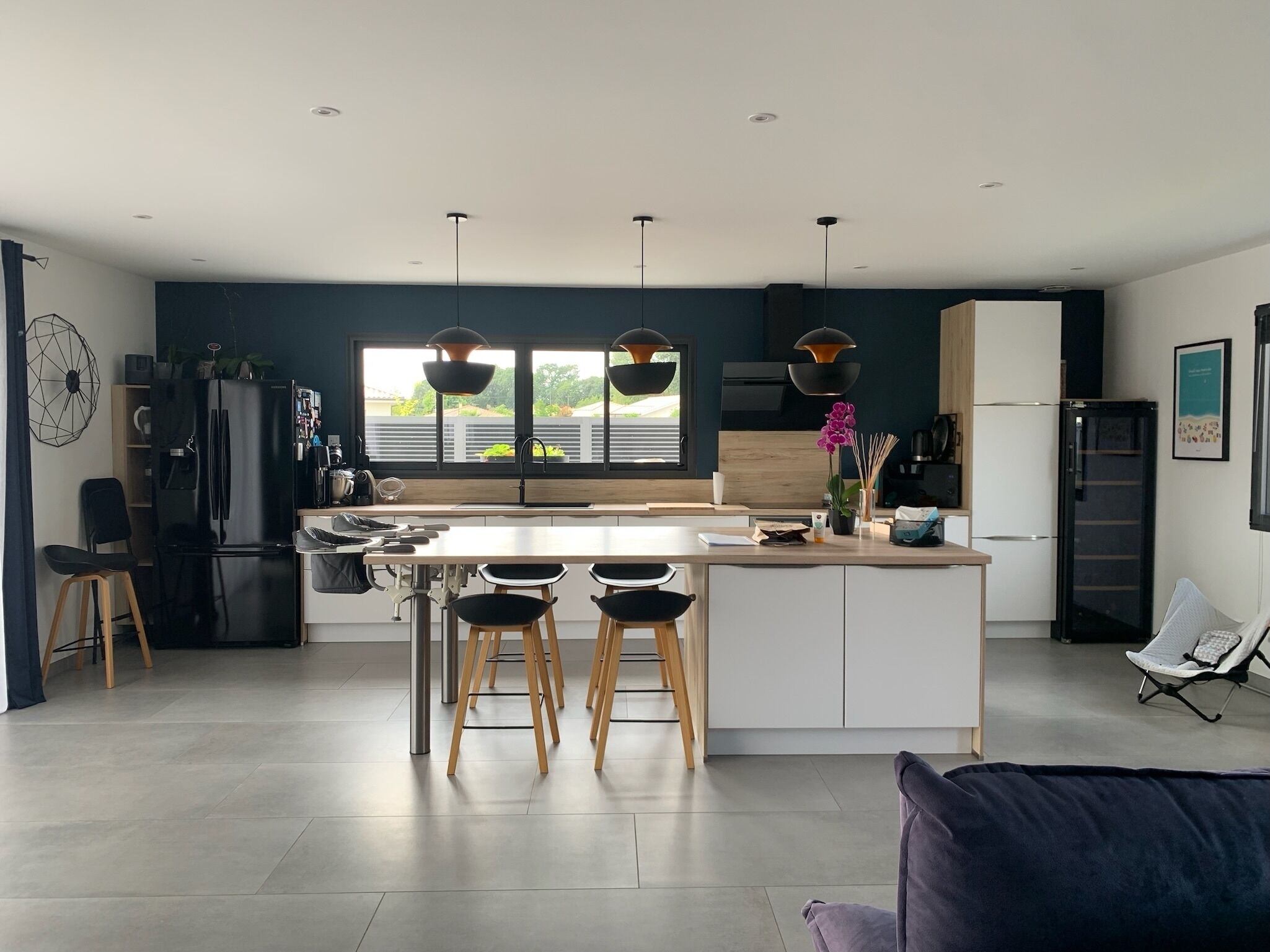 A modern kitchen with a central island, pendant lights, and a view of greenery outside.