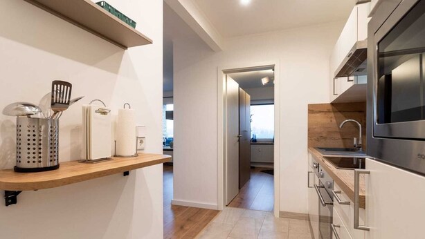 Cuina moderna amb una prestatgeria de fusta, electrodomèstics d'acer inoxidable i vista cap a una habitació veïna.