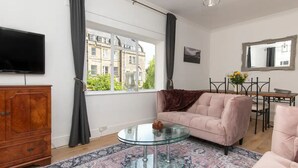 Ein Wohnzimmer mit einer Couch, einem Glastisch, einem Holzschrank, einem Fernseher und Blick auf Gebäude durch große Fenster.