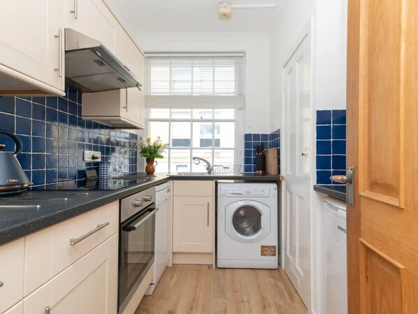 A compact kitchen with a dishwasher, oven, and a window with blinds.