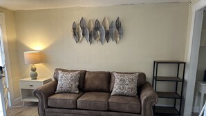 Ein braunes Ledersofa mit gemusterten Kissen, ein kleiner weißer Beistelltisch mit Lampe und eine wandhängende, dekorative Blatanordnung.