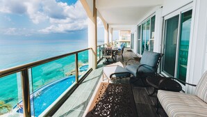 A balcony with a view of the ocean, a pool, and a seating area with a table and chairs.