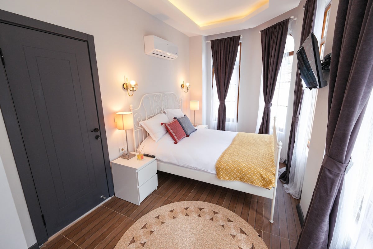 A bedroom with a bed, bedside tables, an air conditioner, curtains, and a television.