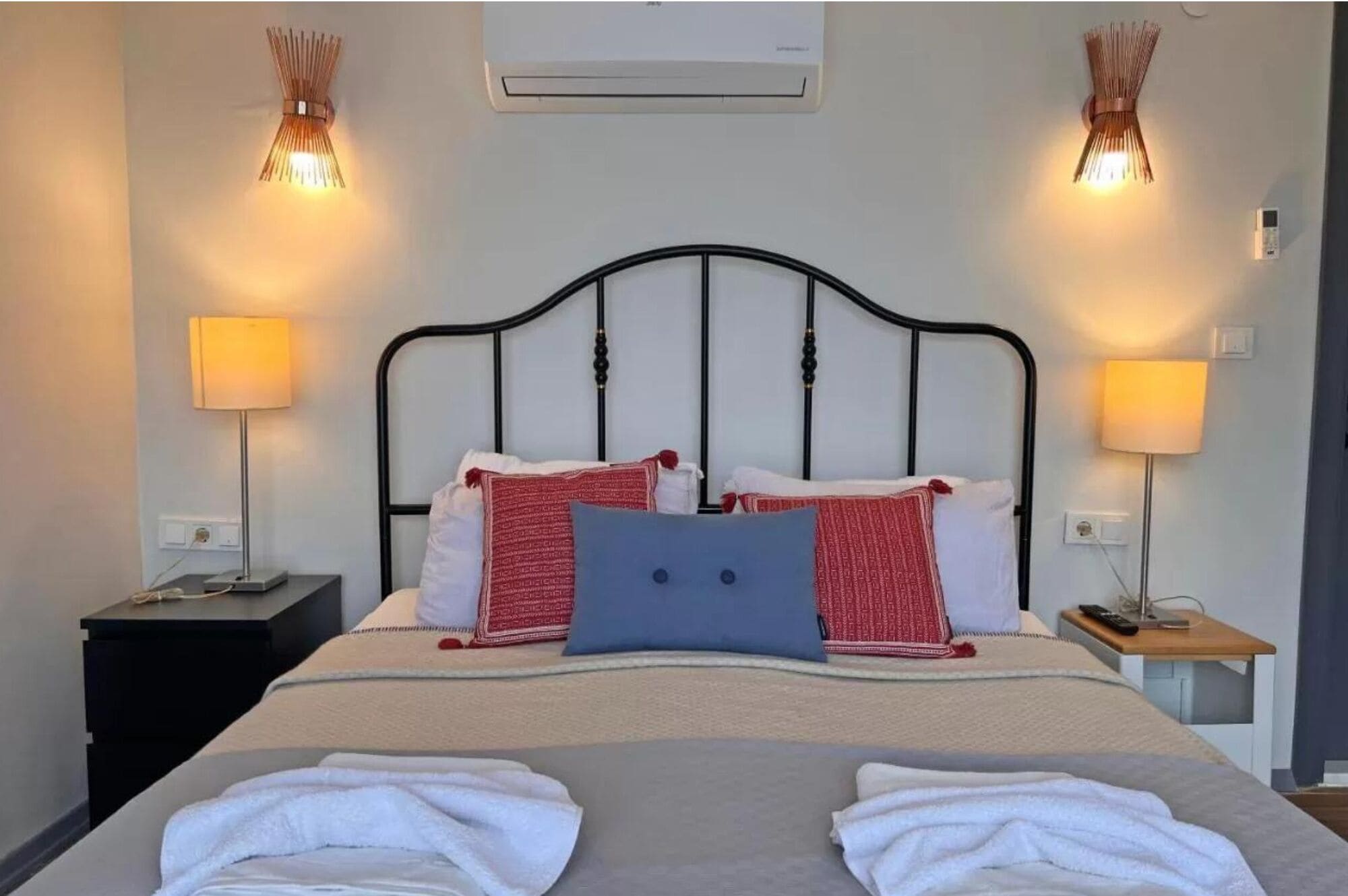 A neatly made bed with a headboard, two bedside tables with lamps, and an air conditioning unit on the wall.