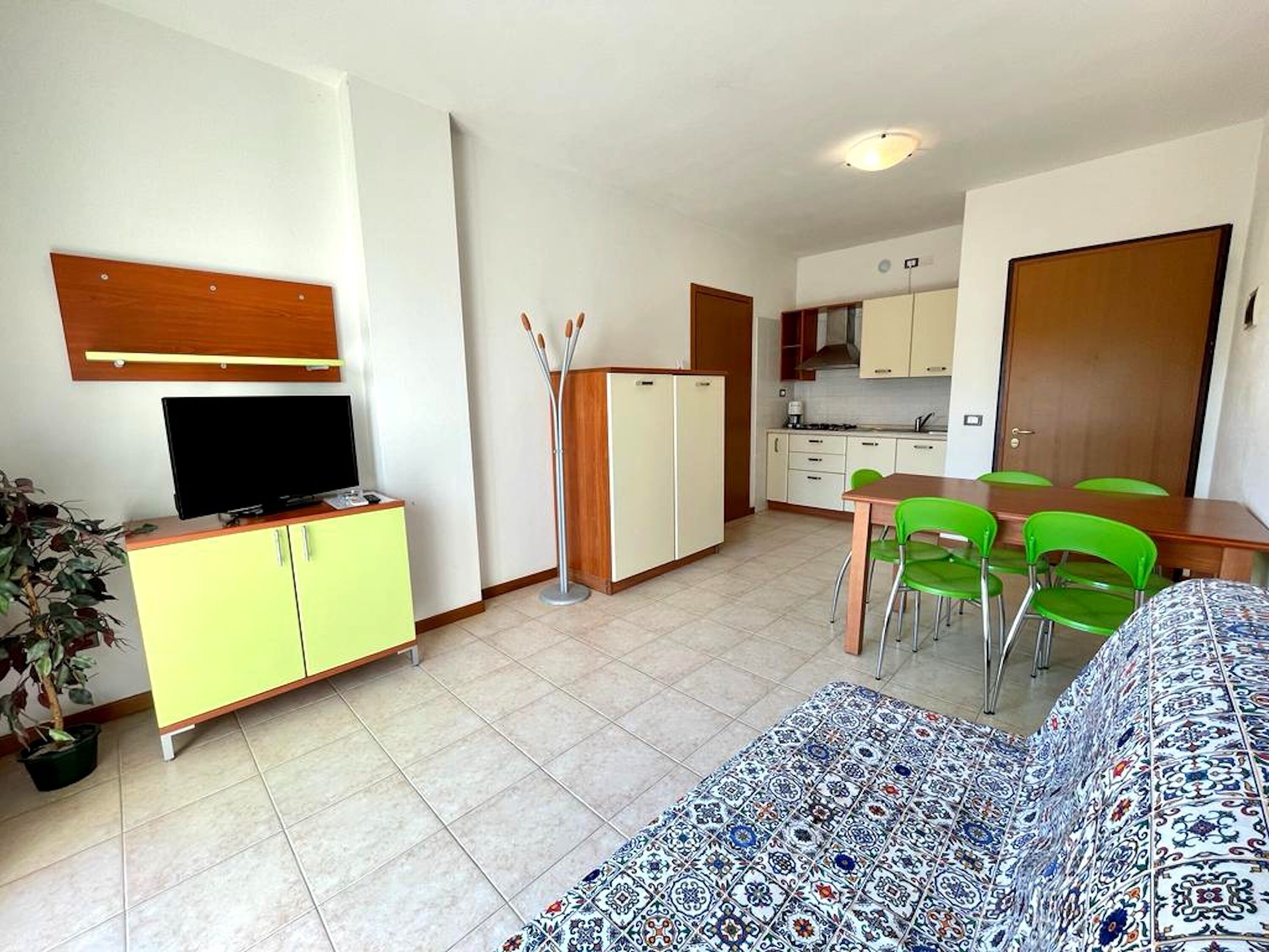 A compact living space with a sofa, a television on a cabinet, a dining table with chairs, and a kitchen area.