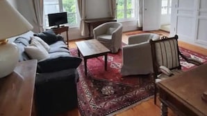 A spacious living room with a sofa set, armchairs, a coffee table, and a wooden dining table. Large windows with curtains allow natural light to fill the room. - Cabourg Plein Centre - Appartement Douillet et Lumineux (Cabourg)