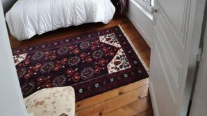 A small bedroom with a bed, a nightstand, a patterned rug, and a window with curtains. - Cabourg Plein Centre - Appartement Douillet et Lumineux (Cabourg)