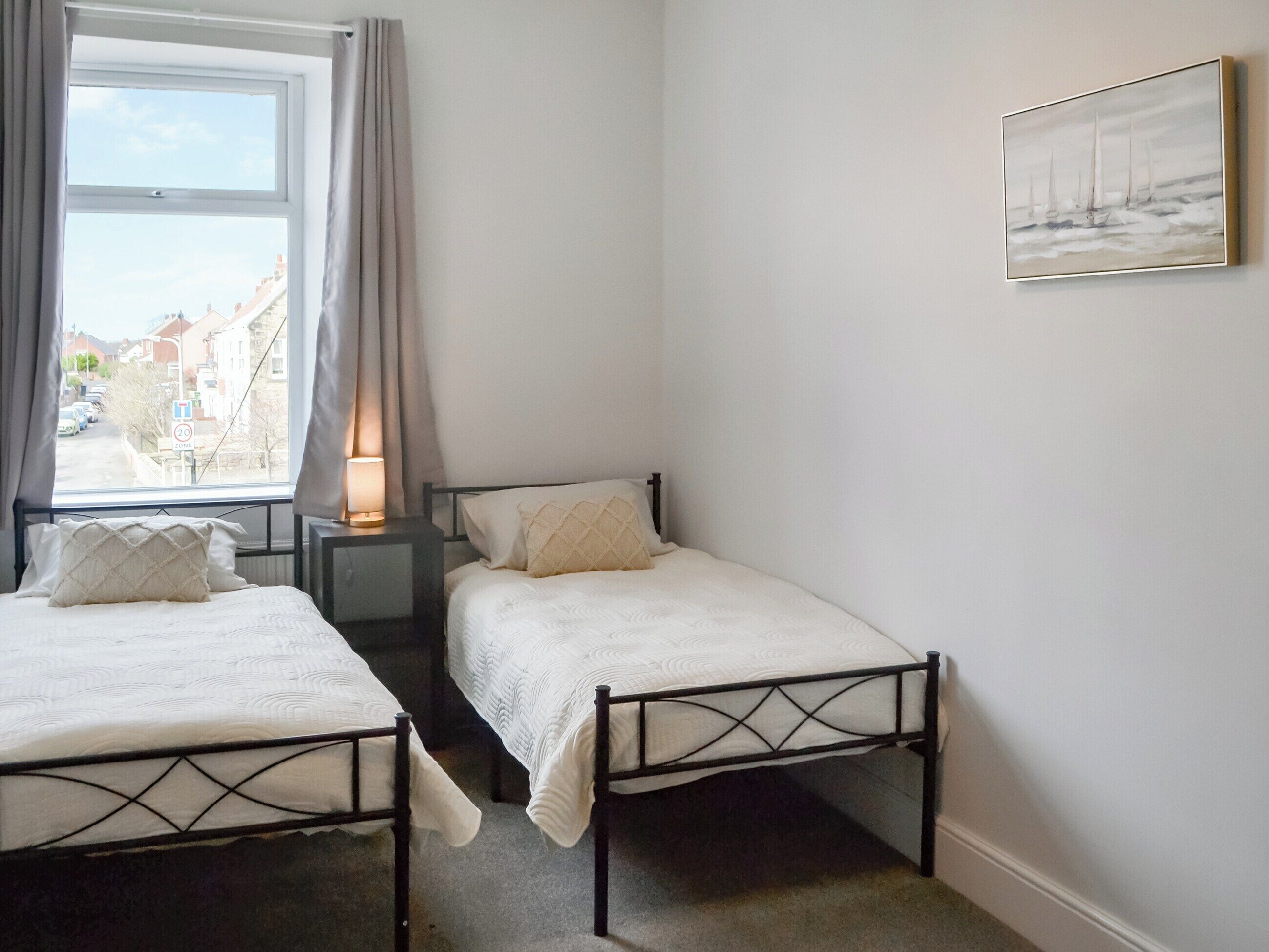 Un dormitorio con dos camas individuales, una mesita de noche con lámpara, una ventana con cortinas y un cuadro en la pared.
