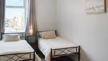 Un dormitorio con dos camas individuales, una mesita de noche con lámpara, una ventana con cortinas y un cuadro en la pared.