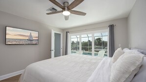 A bedroom with a large bed, a ceiling fan, a window with curtains, and a painting of a sailboat on the wall.