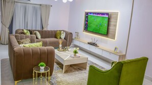 A modern living room with a brown sofa, a white coffee table, and a green armchair. A flat-screen TV is mounted on the wall displaying a sports game. - Luxenest T1 (Lekki)