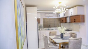 A modern kitchen with white cabinets, a dining area with a table and chairs, and a decorative plant. - Luxenest T1 (Lekki)