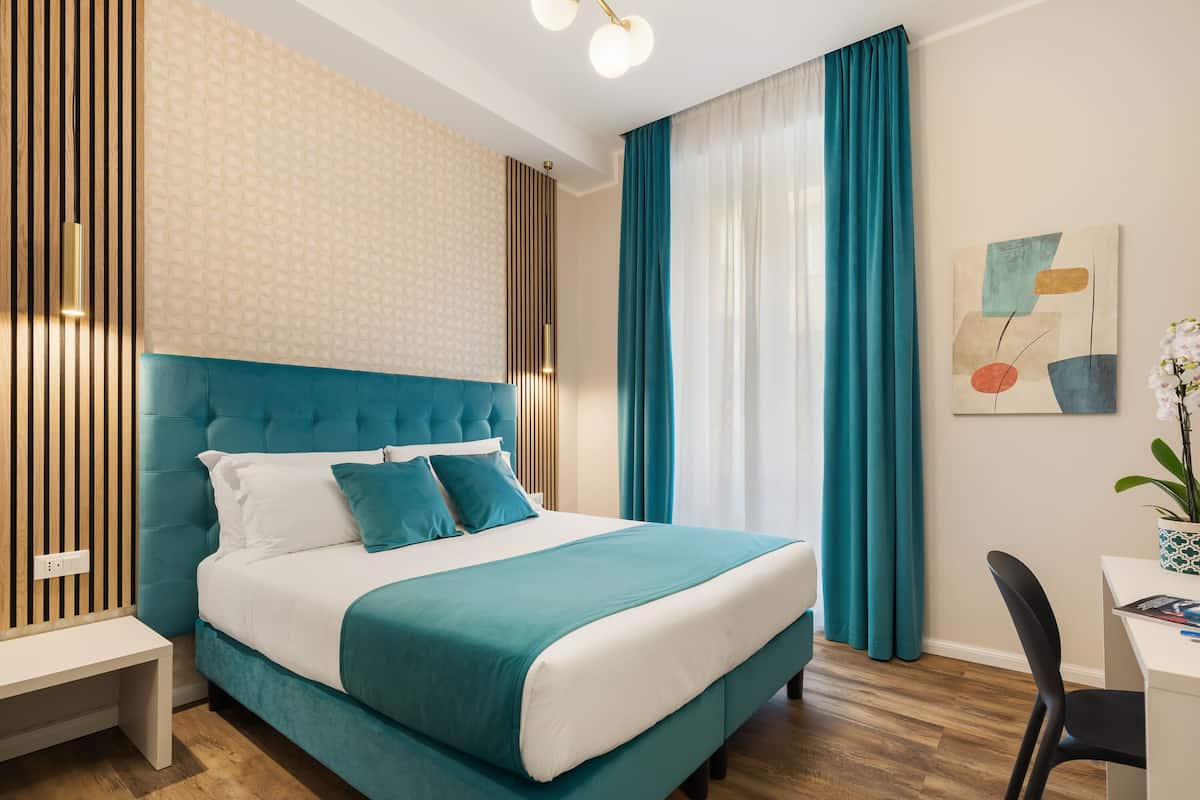 Una habitación de hotel moderna con una cama tapizada en color aguamarina, una mesita de noche blanca y un escritorio con una silla.