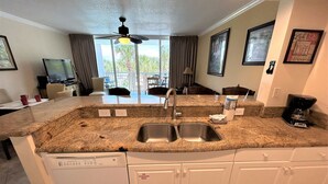 A kitchen with a granite countertop, stainless steel sink, and white cabinetry. There is a ceiling fan, a window with curtains, and a dining area visible in the background.