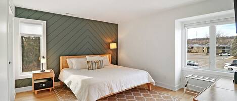 A bedroom with a bed, a nightstand, a lamp, a window, and a view of the outside.