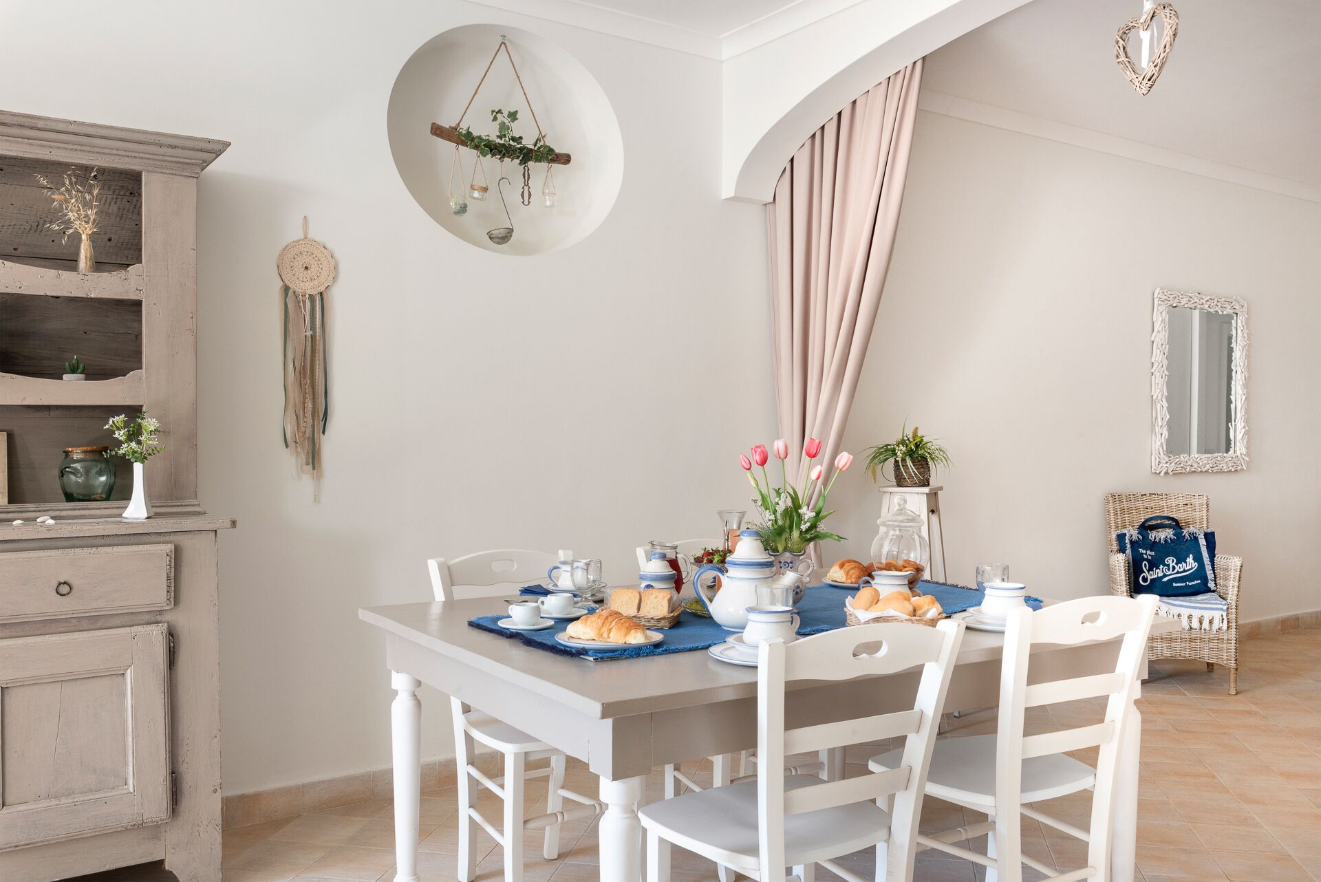 A dining area with a white table set for breakfast, including croissants and a teapot. There are chairs around the table, and a wicker chair is visible in the background.