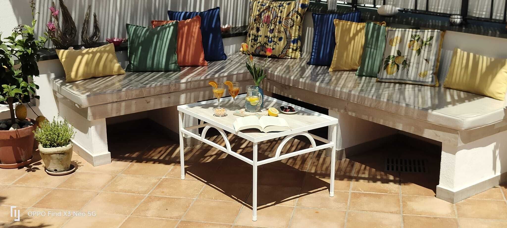 Ein Sitzbereich im Freien mit einem weißen Metalltisch, einer Bank mit bunten Kissen und Topfpflanzen.