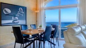 Un coin repas avec une table dressée pour quatre personnes, une grande œuvre d’art encadrée au mur et une vue sur la mer par une grande fenêtre en arc de cercle.