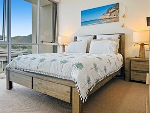 A bedroom with a bed, nightstands, a lamp, and a painting of a beach. - 1 Bright Point Apartment 1407 (Nelly Bay)