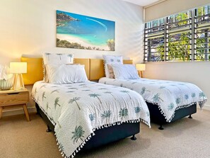 Une chambre avec deux lits, une table de chevet, une lampe, un tableau représentant une plage et une fenêtre donnant sur des palmiers.