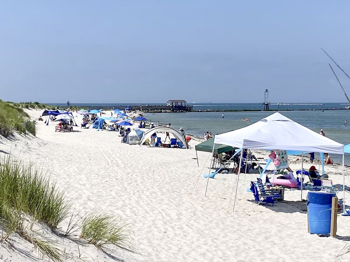 A beach with tents and beach chairs, a pier, and a lighthouse in the background.