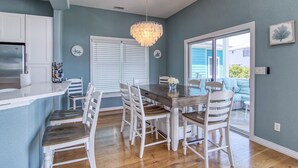 Comedor con mesa de madera y sillas blancas, una araña y vista a una casa azul a través de un vidrio.