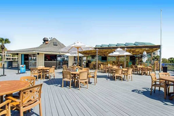 An outdoor deck with wooden tables and chairs, umbrellas, and a building with a sloped roof in the background.