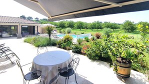 Un espace extérieur aménagé en terrasse, avec une table ronde et des chaises, un coin salon abrité et une vue sur une piscine et un jardin.