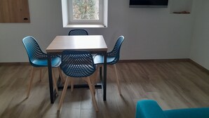 Ein Zimmer mit einem Holztisch und vier blauen Stühlen, einem Fenster mit Blick auf Bäume, einem flachbildfernseher an der Wand und einer Holztür.