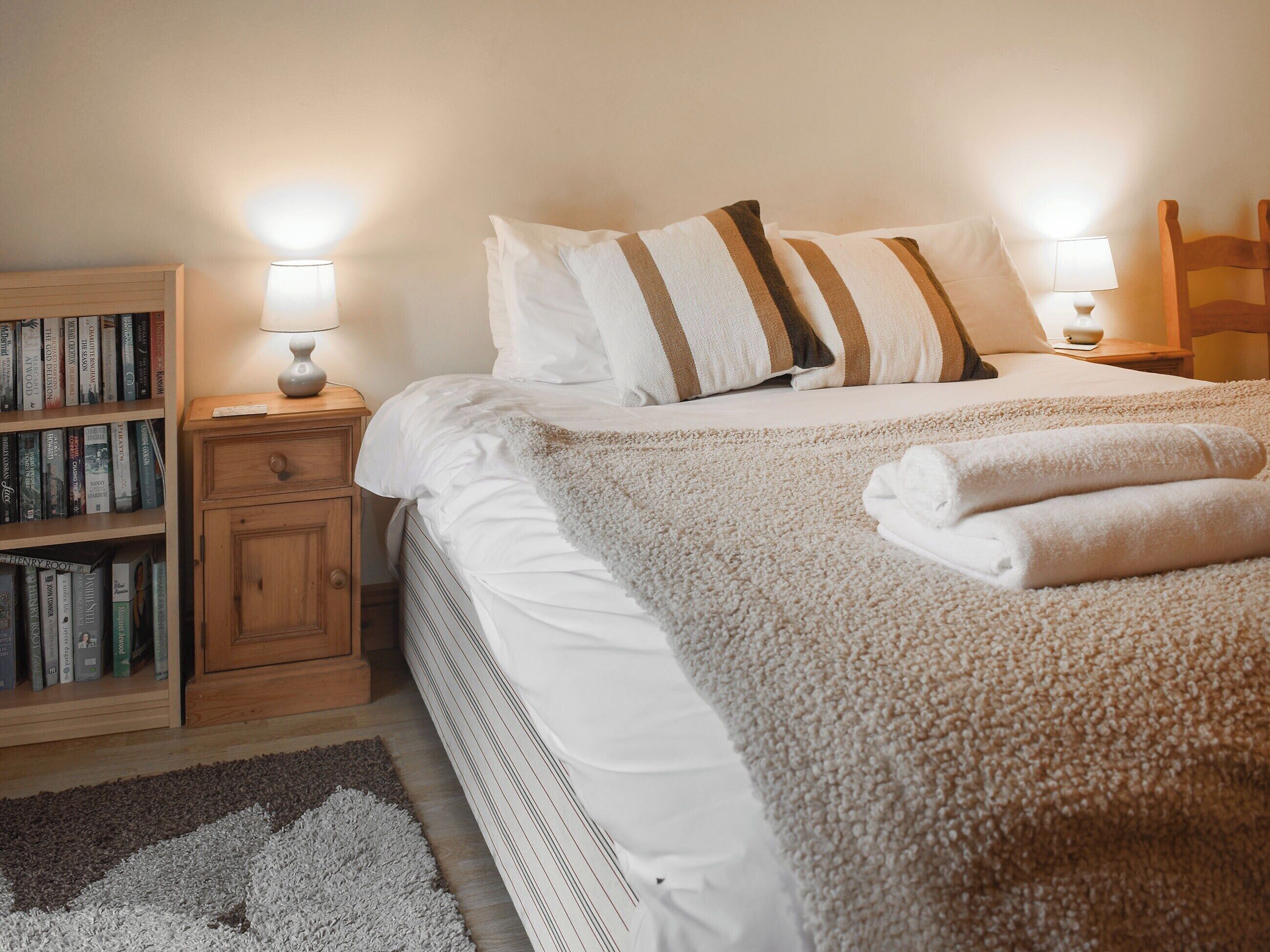 Una cama bien tendida con sábanas blancas y almohadas a rayas, una mesita de noche de madera con una lámpara, y una estantería llena de libros.